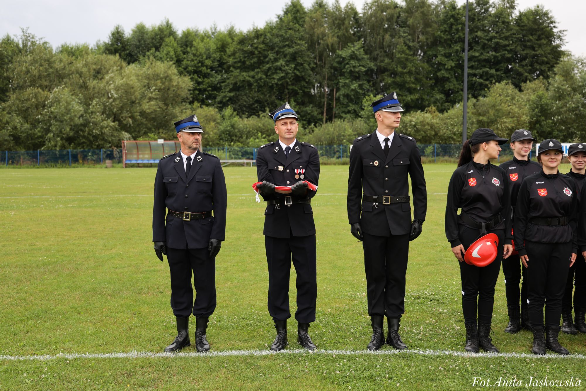 Grupa umundurowanych strażaków na zielonym boisku, trzech mężczyzn w galowych mundurach z czarnymi rękawiczkami, kobieta w czarnym stroju trzyma czerwoną hełmę.