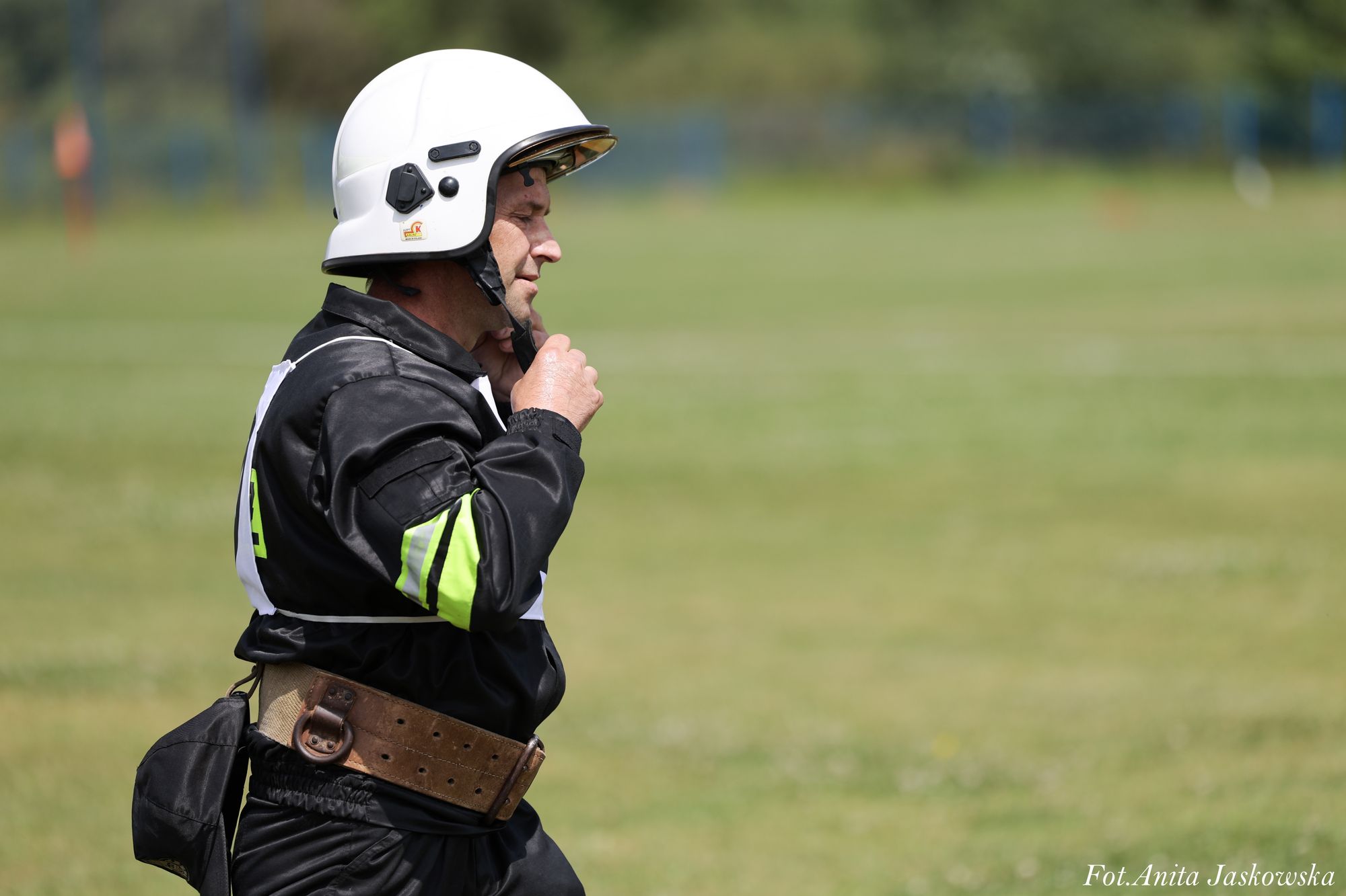 Mężczyzna w czarnym kombinezonie z odblaskowymi paskami i białym kaskiem zapinający pasek pod brodą, na tle zielonej łąki.