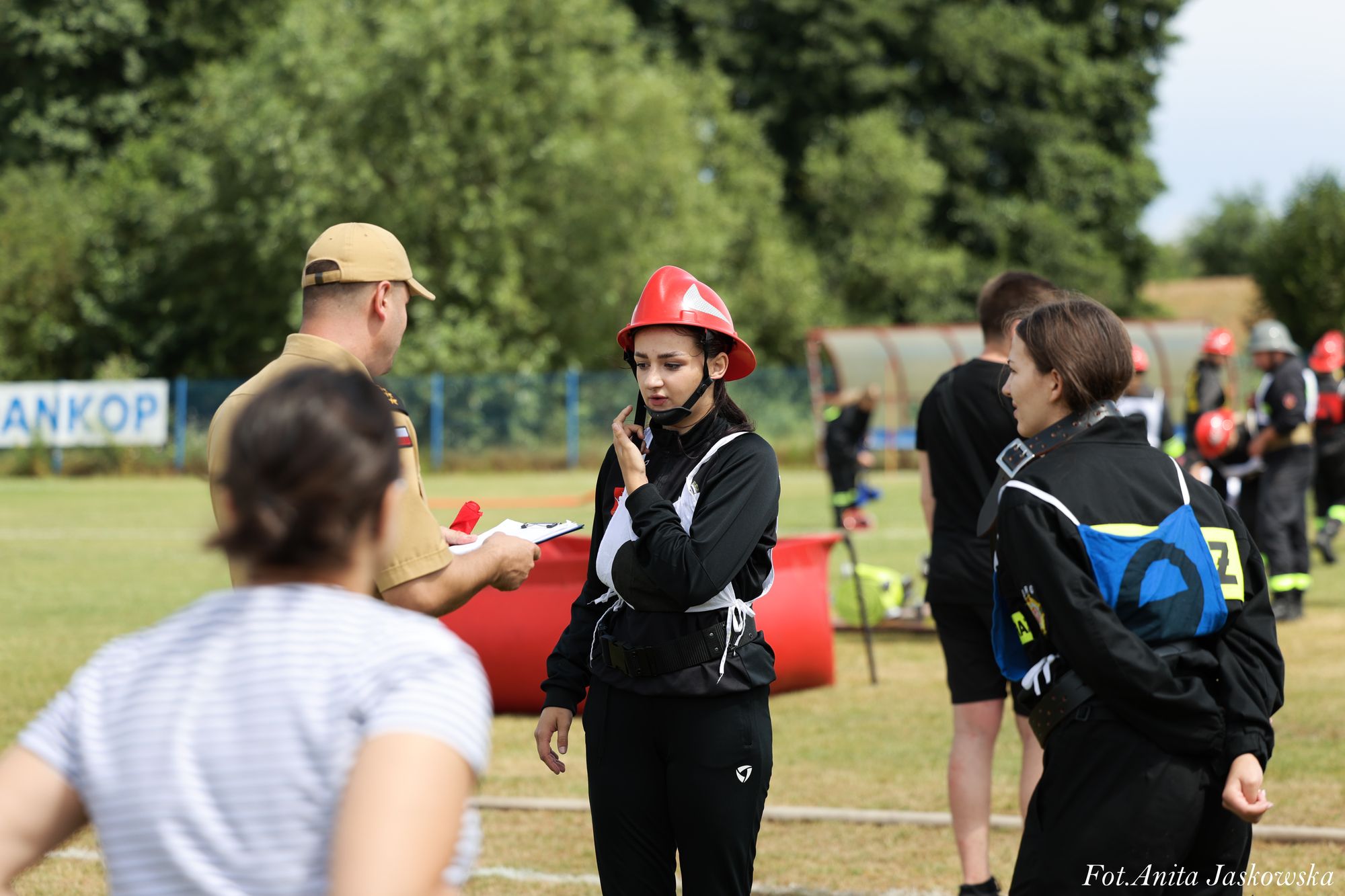 Kobieta w czerwonym hełmie i czarnym stroju strażaka rozmawia z mężczyzną w beżowym mundurze, na tle zielonego pola i innych osób w strojach strażackich.