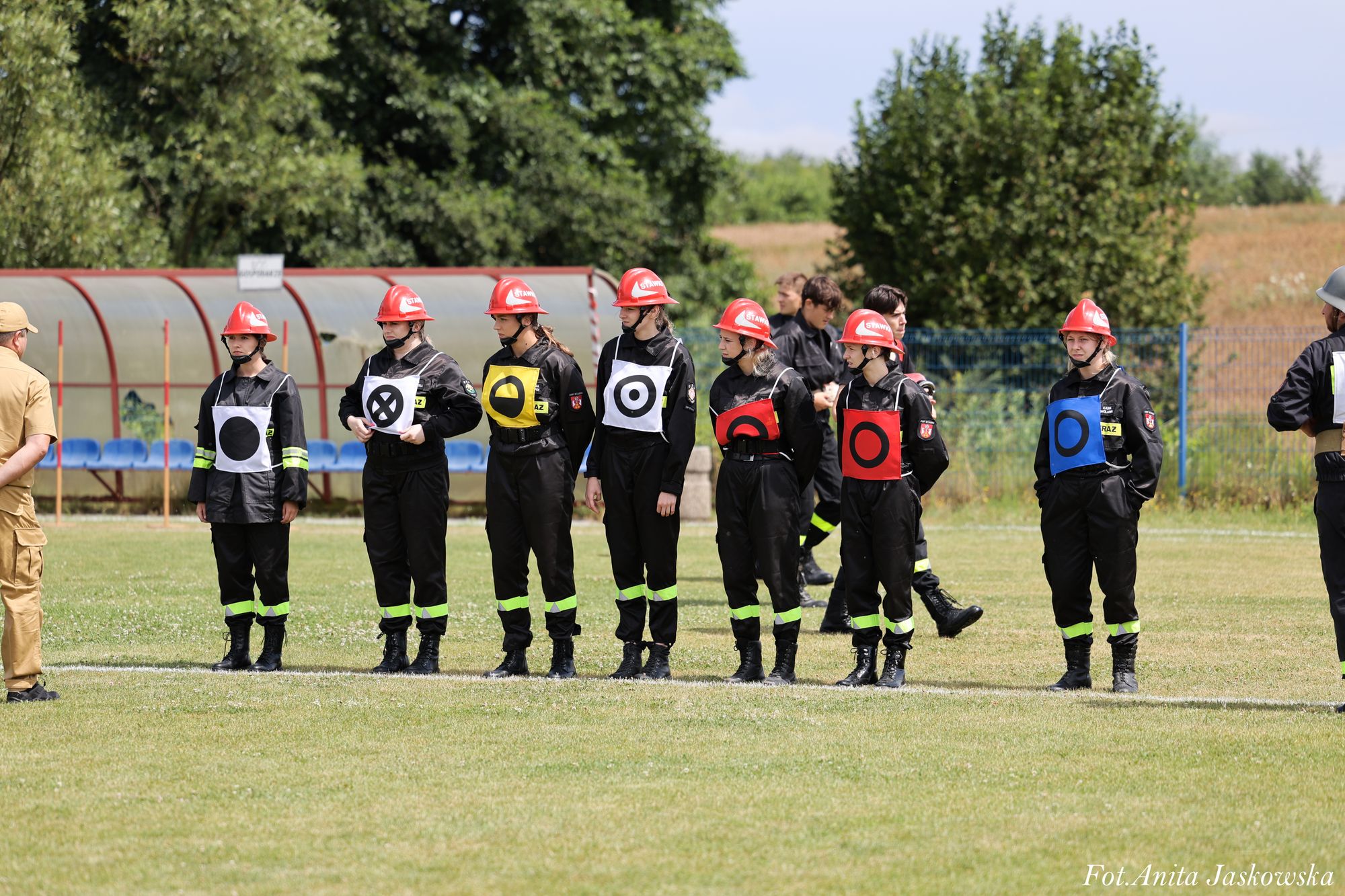 Siedem osób w czarnych mundurach i czerwonych hełmach stoi na trawie, każda z kolorowym kwadratowym oznaczeniem z czarnymi symbolami, w tle drzewa i zadaszone ławki.