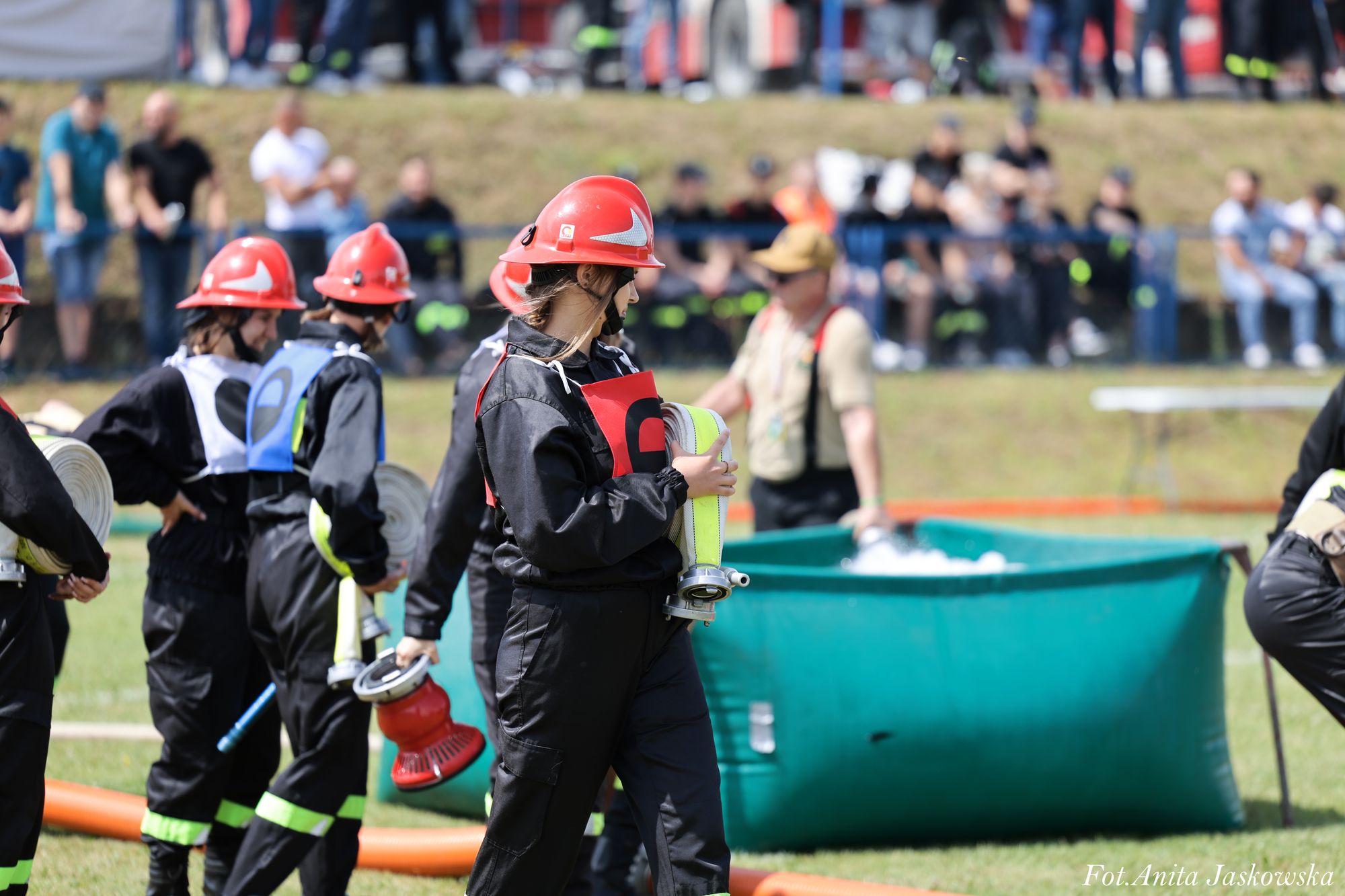 Grupa osób w czarnych ubraniach i czerwonych hełmach, trzymających zrolowane węże strażackie, na trawie przed dużym zielonym zbiornikiem z wodą.