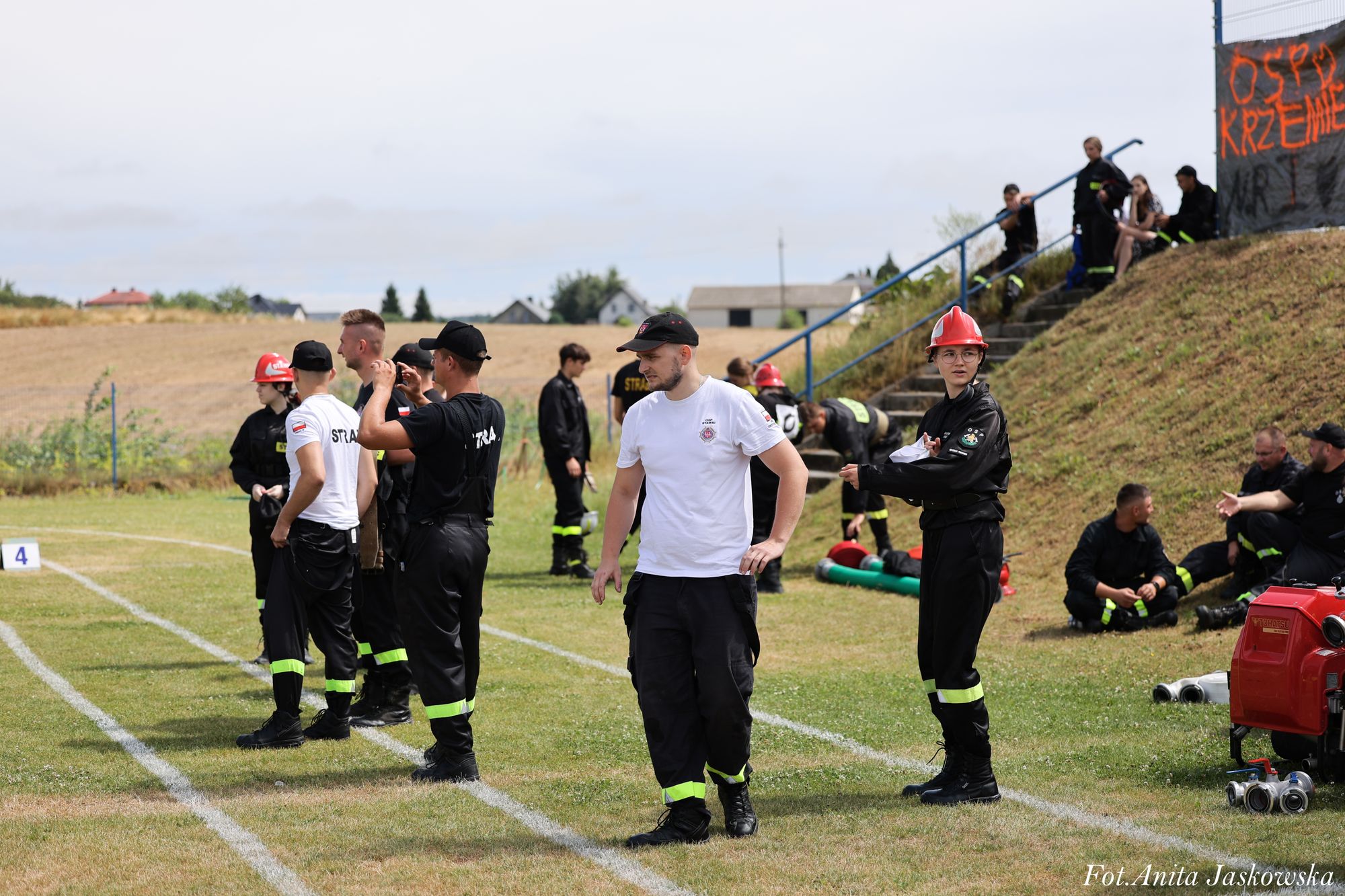 Grupa osób w czarnych i białych mundurach z odblaskowymi pasami na trawie z oznaczonymi liniami, niektórzy w czerwonych hełmach, wokół sprzęt strażacki.