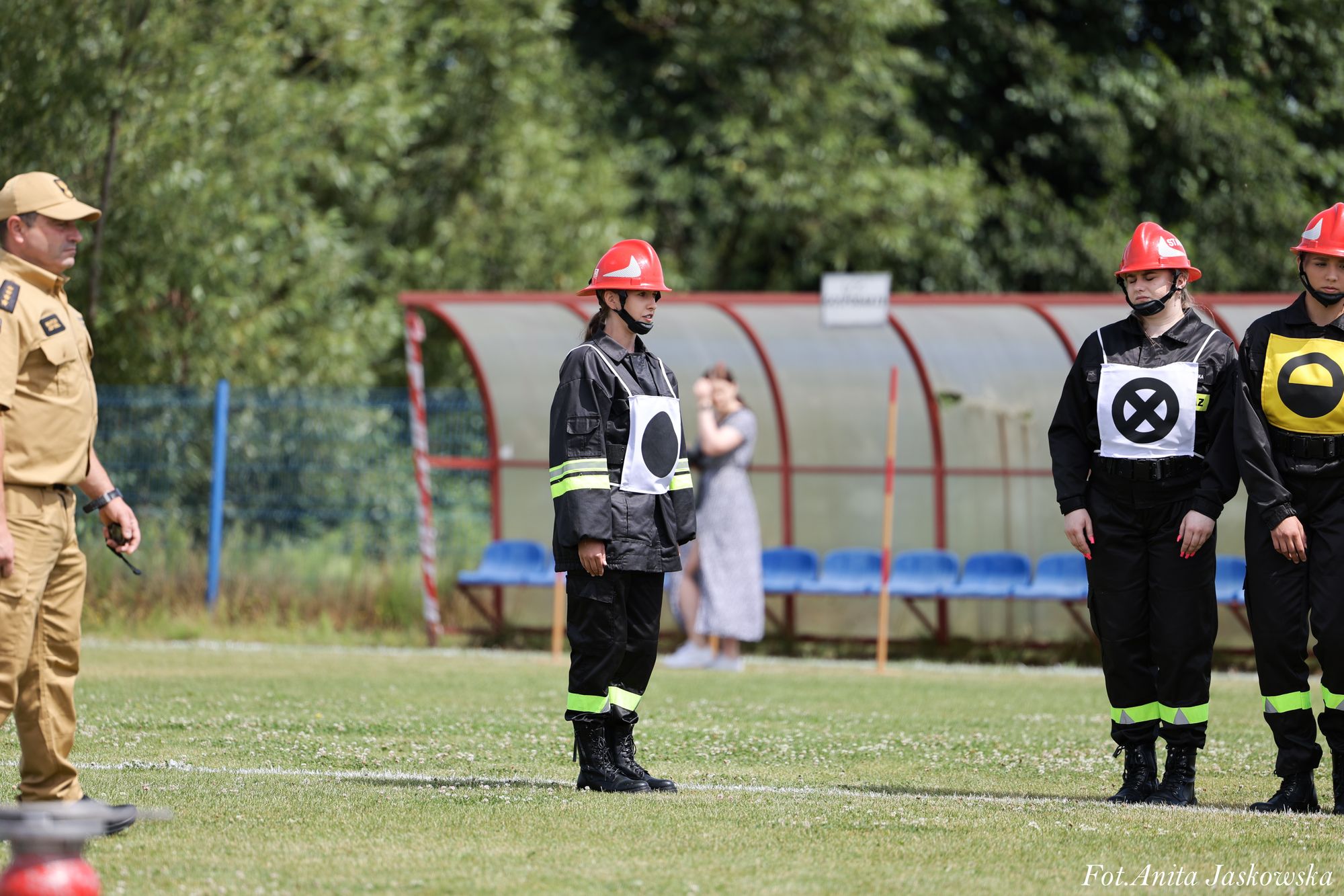 Trzy osoby w czarnych mundurach z odblaskowymi paskami i czerwonych hełmach, jedna w beżowym mundurze, stoją na zielonym boisku sportowym, w tle ławki i zarośla.