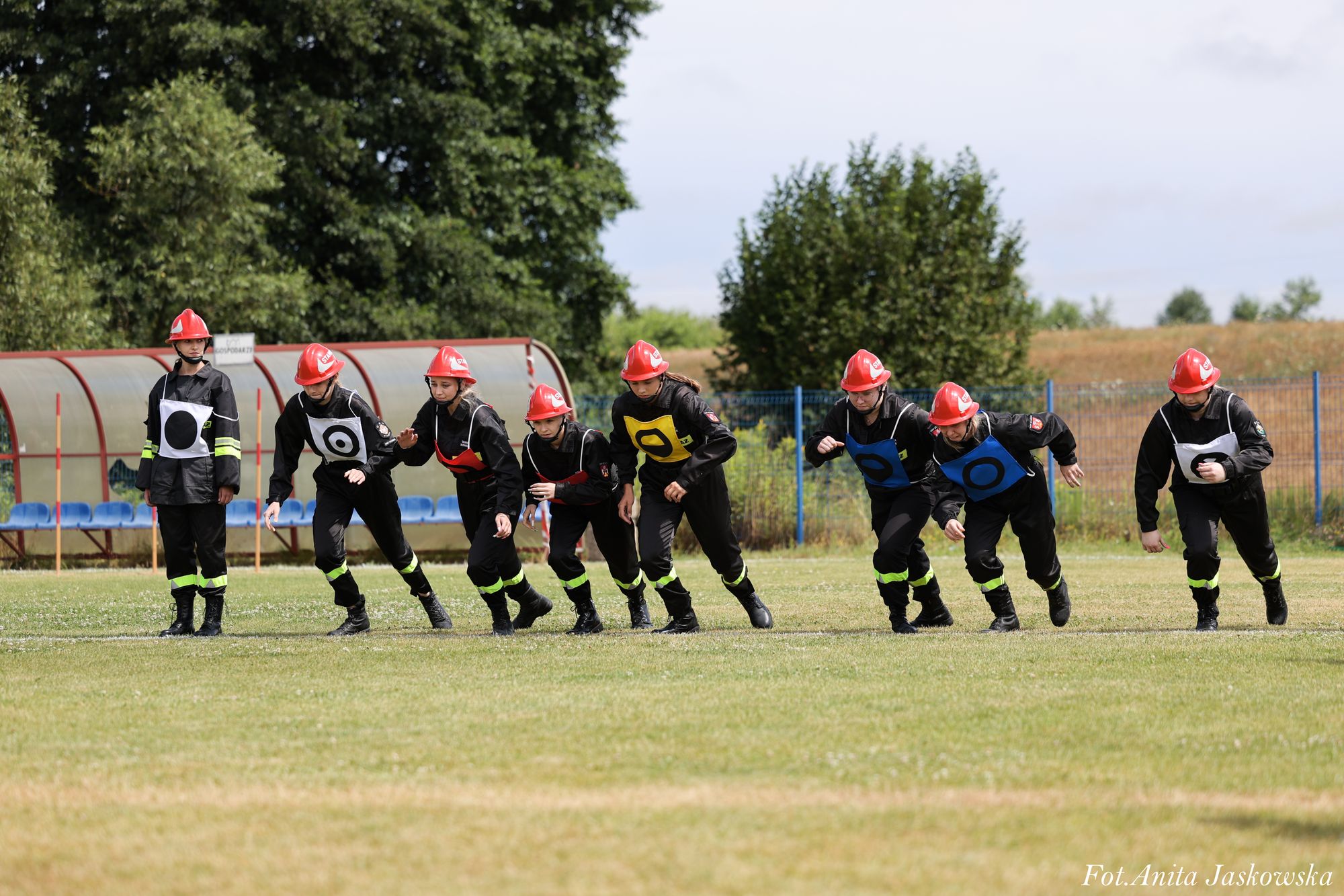 Osoby w czarnych mundurach z czerwonymi hełmami, na trawie, ustawione w linii, niektóre w trakcie startu do biegu, tło z drzewami i niebieskimi siedzeniami.
