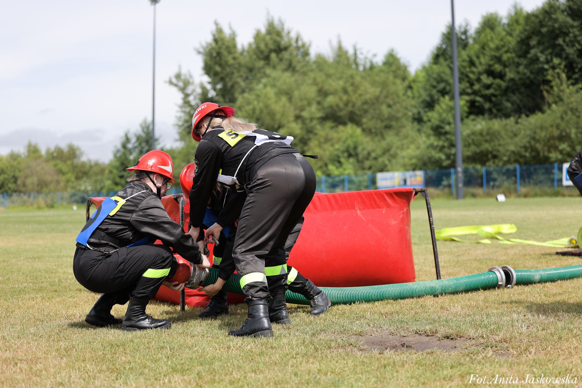 Trzy osoby w czarnych strojach z odblaskowymi paskami i czerwonych hełmach manewrują zielonym wężem na trawiastym boisku, obok czerwony pojemnik.