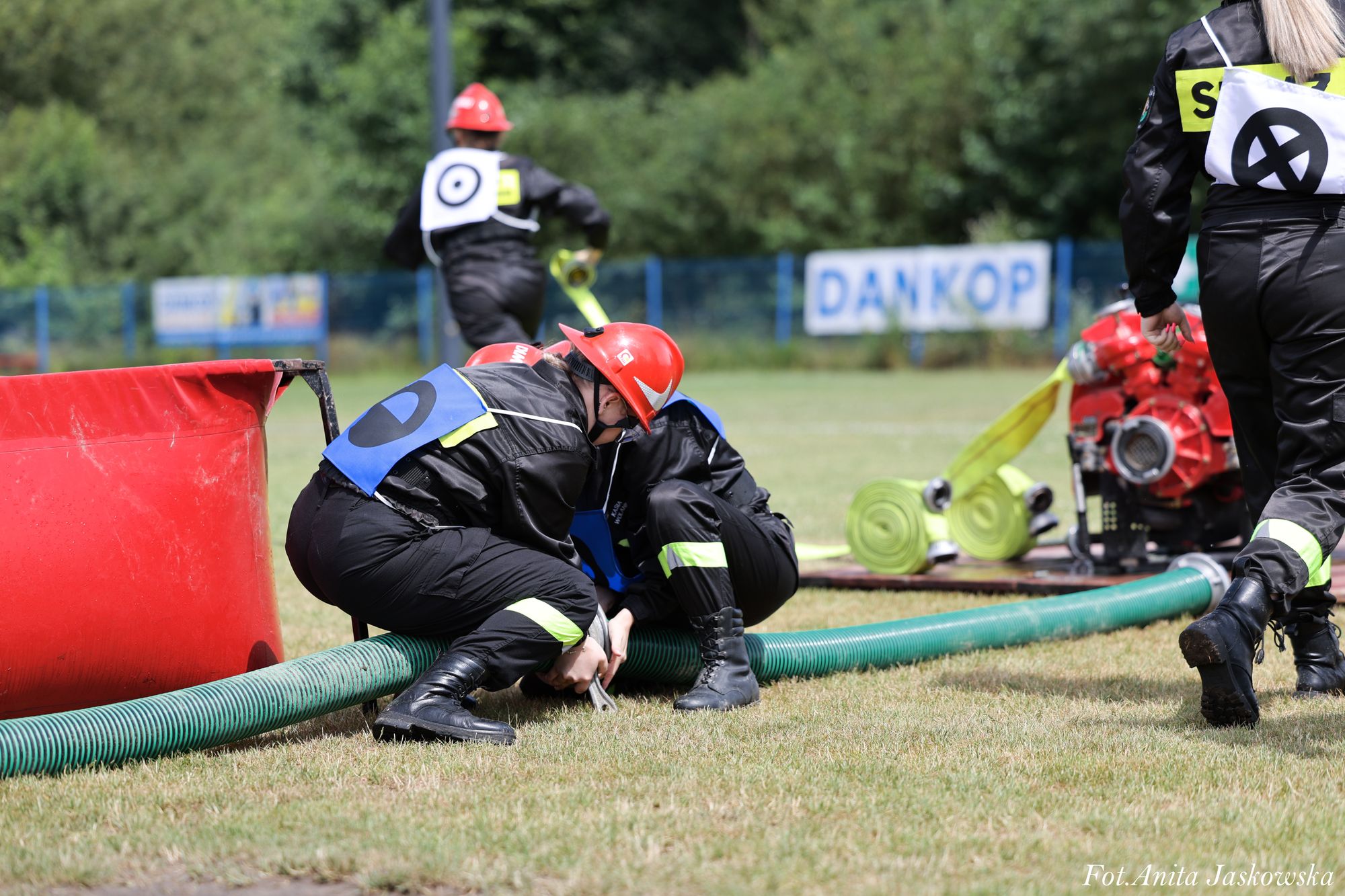 Cztery osoby w czarnych mundurach i czerwonych hełmach pracują na trawie przy zielonym wężu strażackim i czerwonym zbiorniku. W tle tablica z napisem "DANKOP".