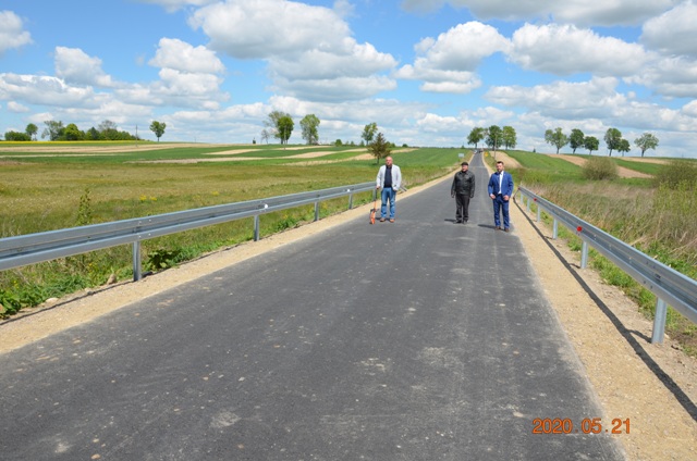 Trzy osoby stoją na szerokiej, nowej asfaltowej drodze z metalowymi barierkami, otoczonej zielonymi polami i rozmytym niebieskim niebem z chmurami.