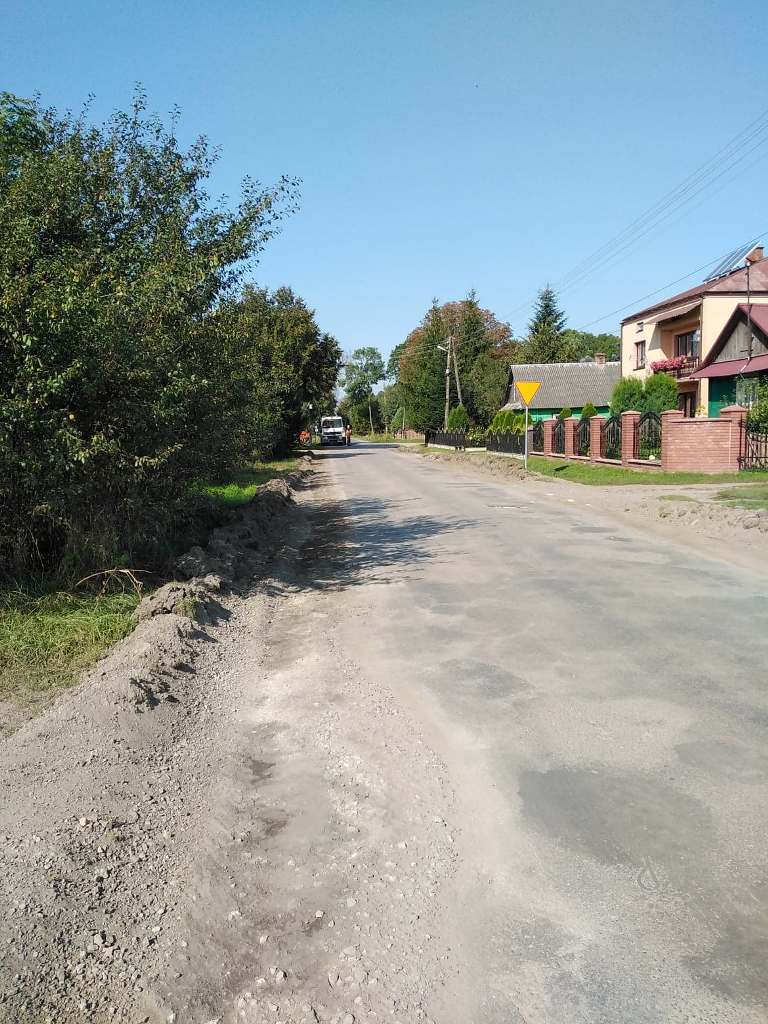 Droga z nierówną, szarą nawierzchnią, po lewej stronie zielone krzewy i drzewa, po prawej czerwony mur i domy, w tle biały samochód i znak drogowy na żółtym tle.