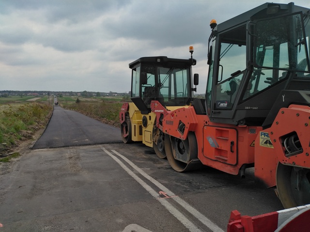 Maszyny drogowe w kolorach pomarańczowym i żółtym stojące obok nowo wyasfaltowanej drogi na tle pochmurnego nieba.