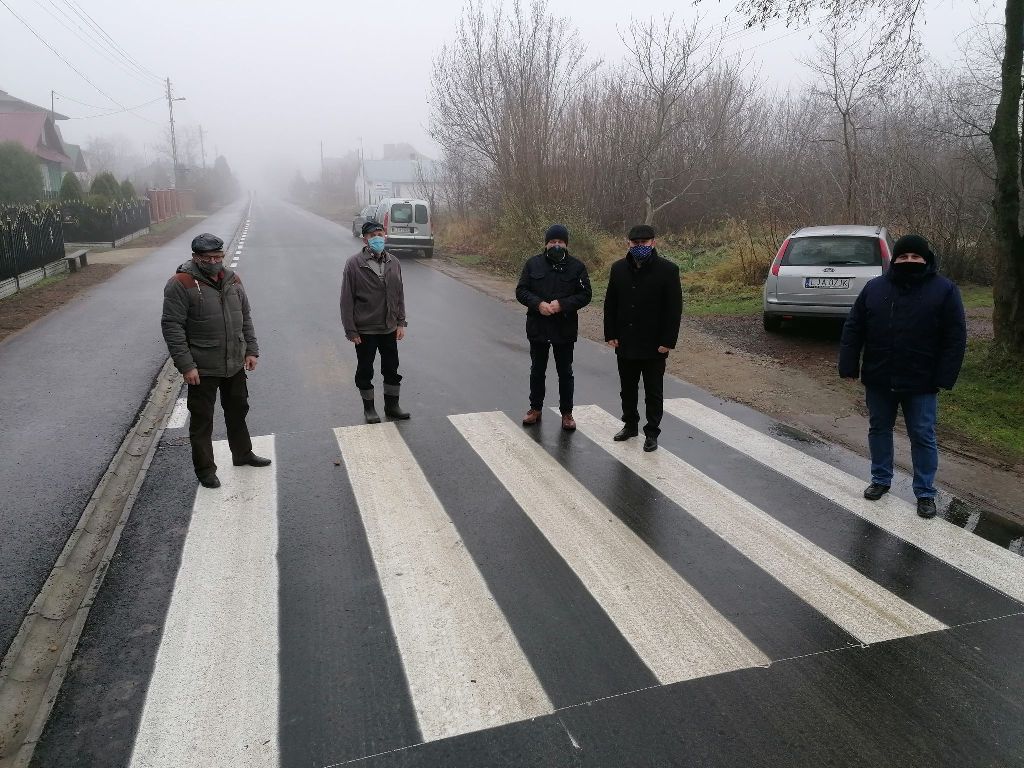 Pięciu mężczyzn w kurtkach i czapkach stoi na przejściu dla pieszych na mokrej, asfaltowej drodze, w tle mgła, drzewa bez liści i zaparkowane samochody.