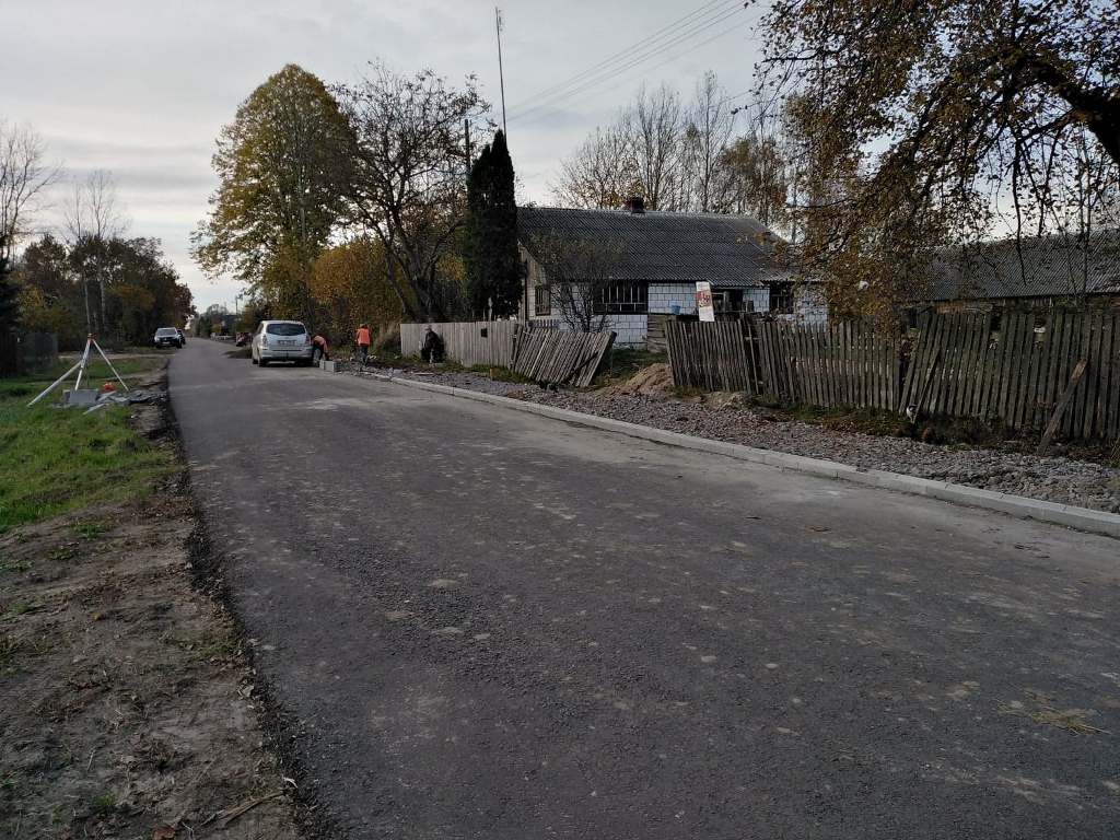 Droga asfaltowa z nowym krawężnikiem, samochody, drewniane, częściowo uszkodzone ogrodzenie, drzewa z jesiennymi liśćmi, szary budynek, pochmurne niebo.