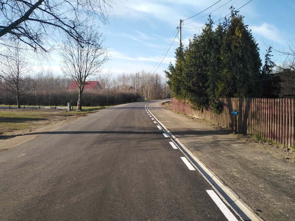 Asfaltowa droga z białymi liniami po prawej stronie, po prawej drewniany płot i zielone drzewa, po lewej trawa, krzewy i dom z czerwonym dachem, niebieskie niebo.