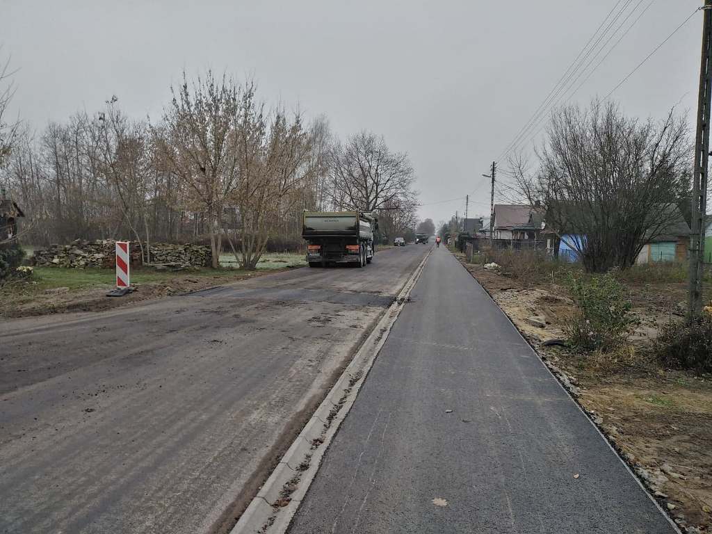 Droga asfaltowa z pasem do jazdy i chodnikiem po prawej stronie, po lewej stronie ciężarówka, drzewa bez liści, pochmurne niebo.