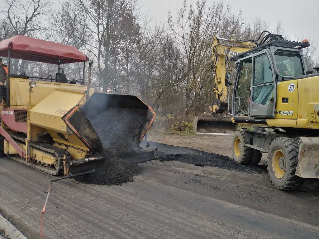 Maszyna do układania asfaltu z wysypującym się gorącym asfaltem na drogę obok żółtego koparko-ładowarki, w tle nagie drzewa i szare niebo.