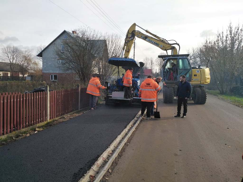 Maszyna budowlana i cztery osoby w pomarańczowych kamizelkach na tle asfaltowej drogi i ogrodzenia, dom i drzewa w tle, częściowo nowo położony czarny asfalt.