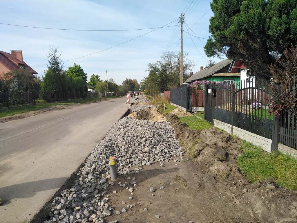 Ulica z wykopanym rowem, częściowo zasypanym kamieniami, po prawej stronie ogrodzenia domów i zielone drzewa, po lewej stronie asfaltowa jezdnia i zieleń.