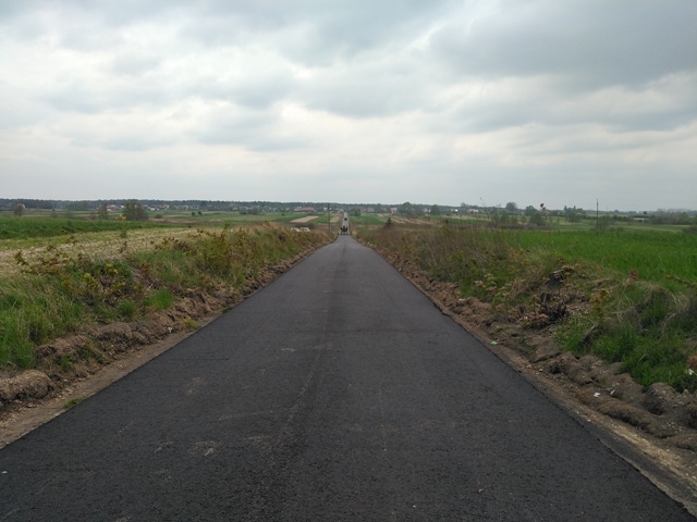 Prosta, świeżo asfaltowana, ciemnoszara droga przez pola pod pochmurnym niebem.