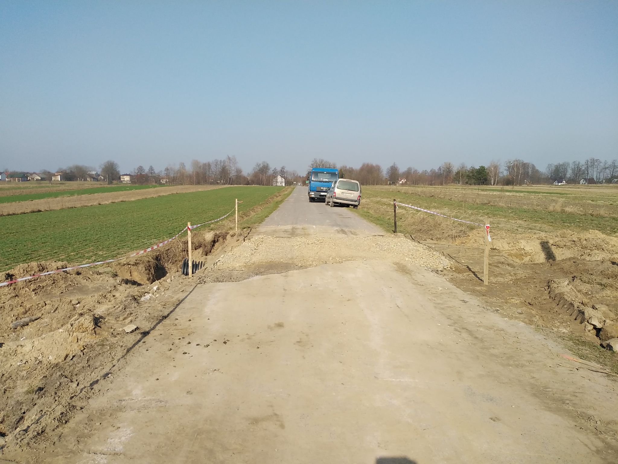Droga gruntowa z wykopem i taśmą ostrzegawczą, na końcu asfaltowa droga z dwoma zaparkowanymi pojazdami, zielone pola i drzewa w tle pod jasnym niebem.