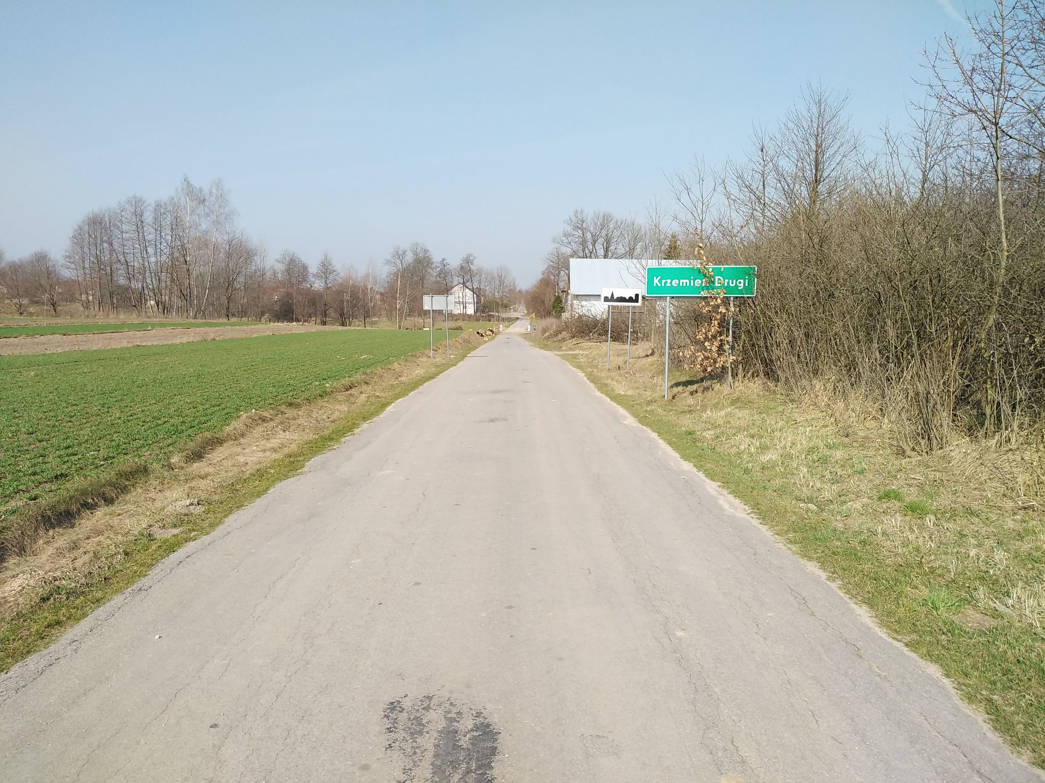 Asfaltowa droga między polami i drzewami, zielona tablica z napisem "Krzemień Drugi" po prawej stronie, bezchmurne niebieskie niebo.