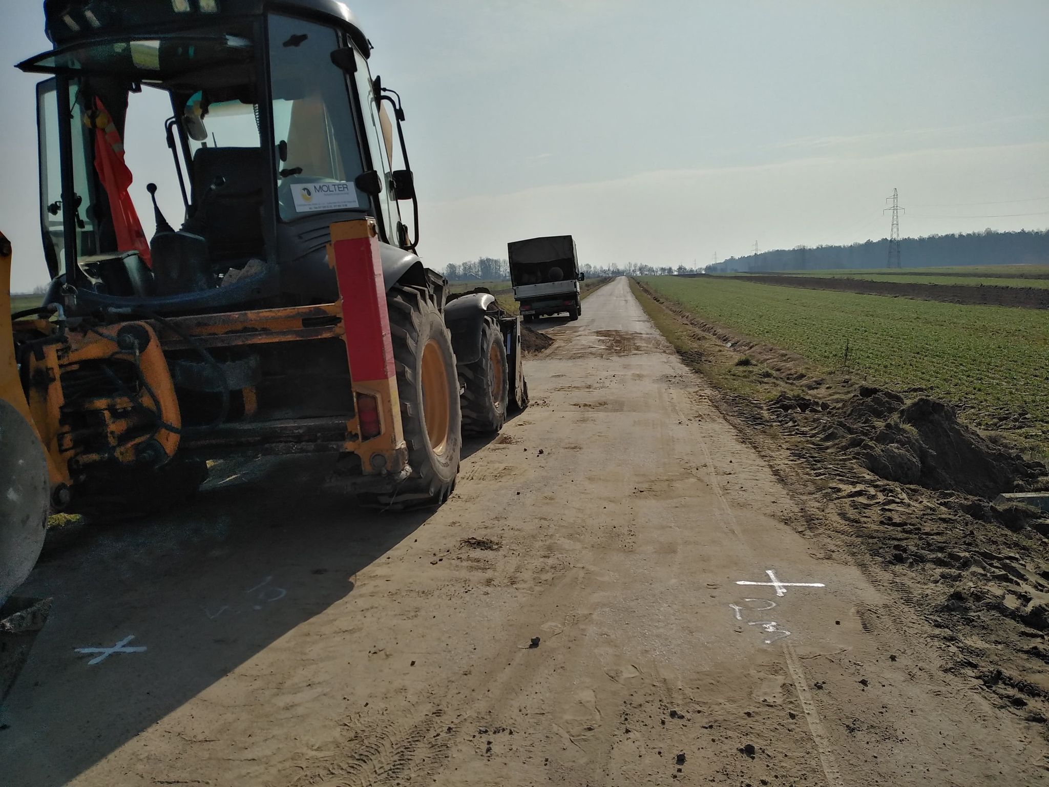 Czarny ciągnik z pomarańczowymi elementami po lewej, droga gruntowa z białymi znakami, zielone pola po prawej, samochód dostawczy na końcu drogi, słupy energetyczne w tle.
