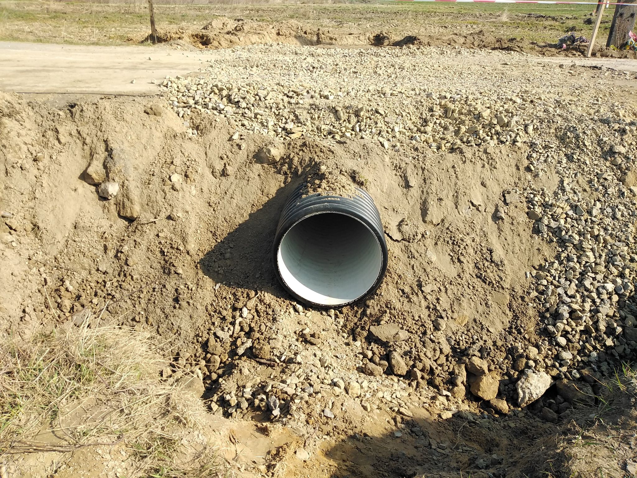 Czarna rura drenarska wkopana w ziemię otoczoną suchą ziemią i żwirem na tle pola.