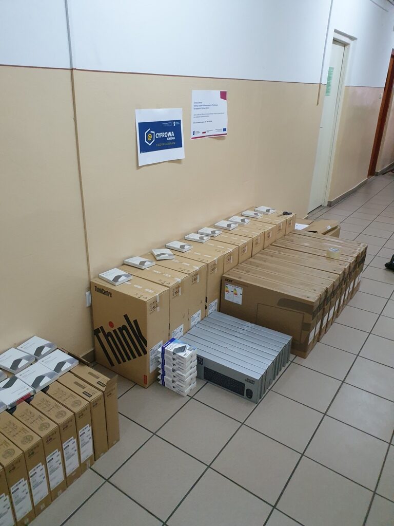 Pudełka komputerów i akcesoriów ułożone na podłodze korytarza z beżowymi ścianami i jasnoszarymi kaflami, na ścianie dwie kartki z nadrukiem.