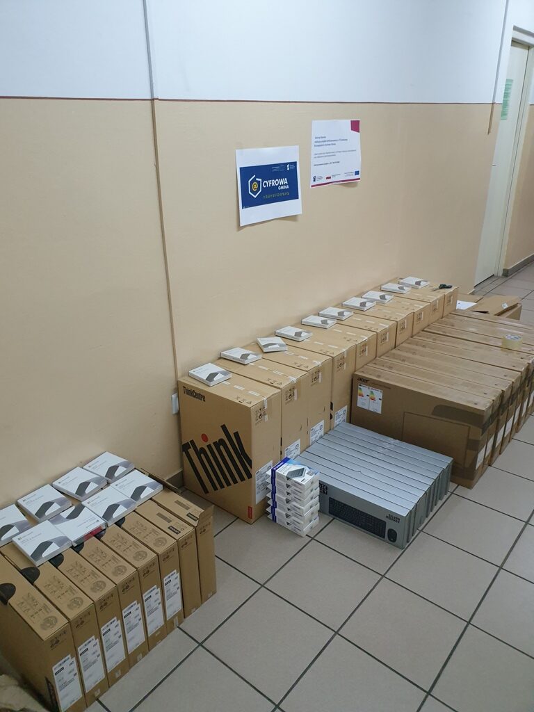 Pudełka z laptopami Lenovo ThinkCentre, klawiatury i inne urządzenia elektroniczne ułożone na beżowej podłodze przy ścianie z kremowo-białą farbą i dwoma plakatami.