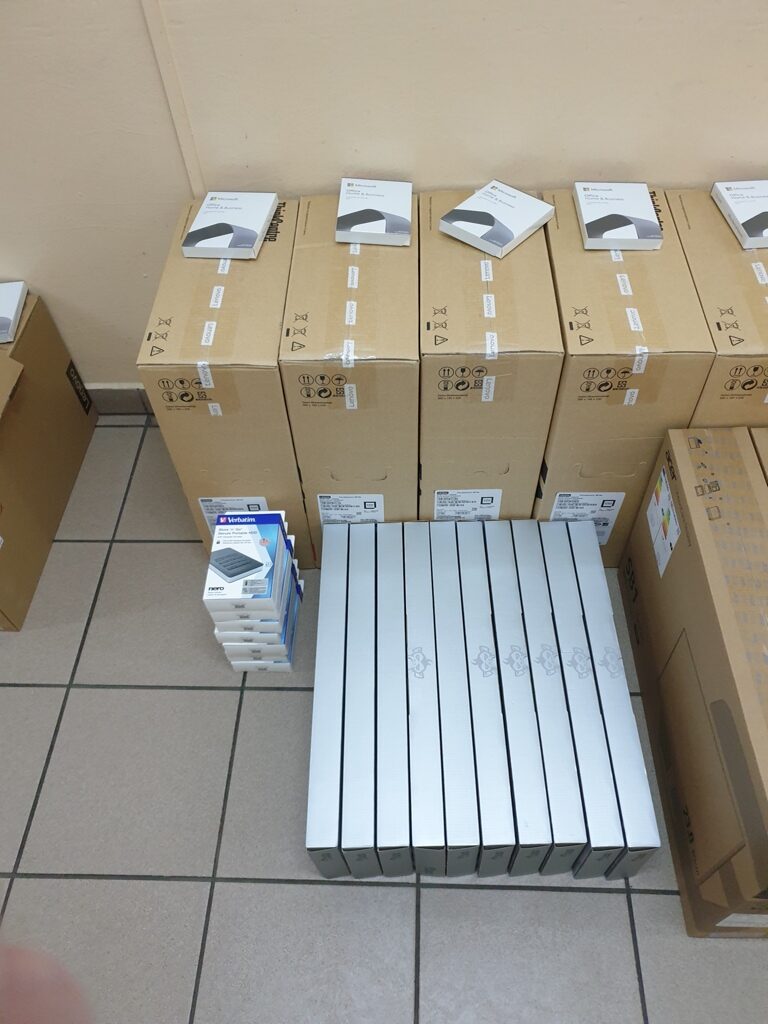 Stosy brązowych kartonów z białymi pudełkami na szczycie oraz rząd białych, długich pudełek i mały stos białych pudełek z niebieskim napisem na jasnej podłodze.