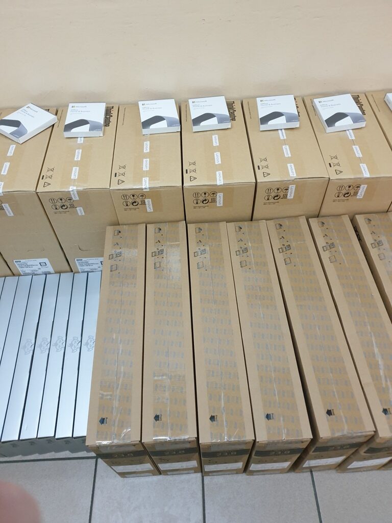 Pudełka kartonowe i białe opakowania ułożone w rzędach na podłodze na tle jasnej ściany.