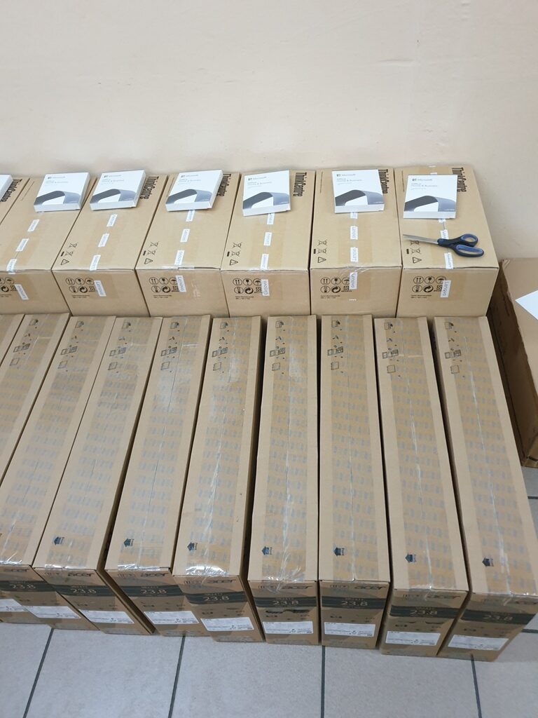 Pudełka w dwóch rzędach, górne mniejsze z białymi etykietami i nożyczkami, dolne większe, prostokątne, brązowe, ułożone na jasnej podłodze przy ścianie.