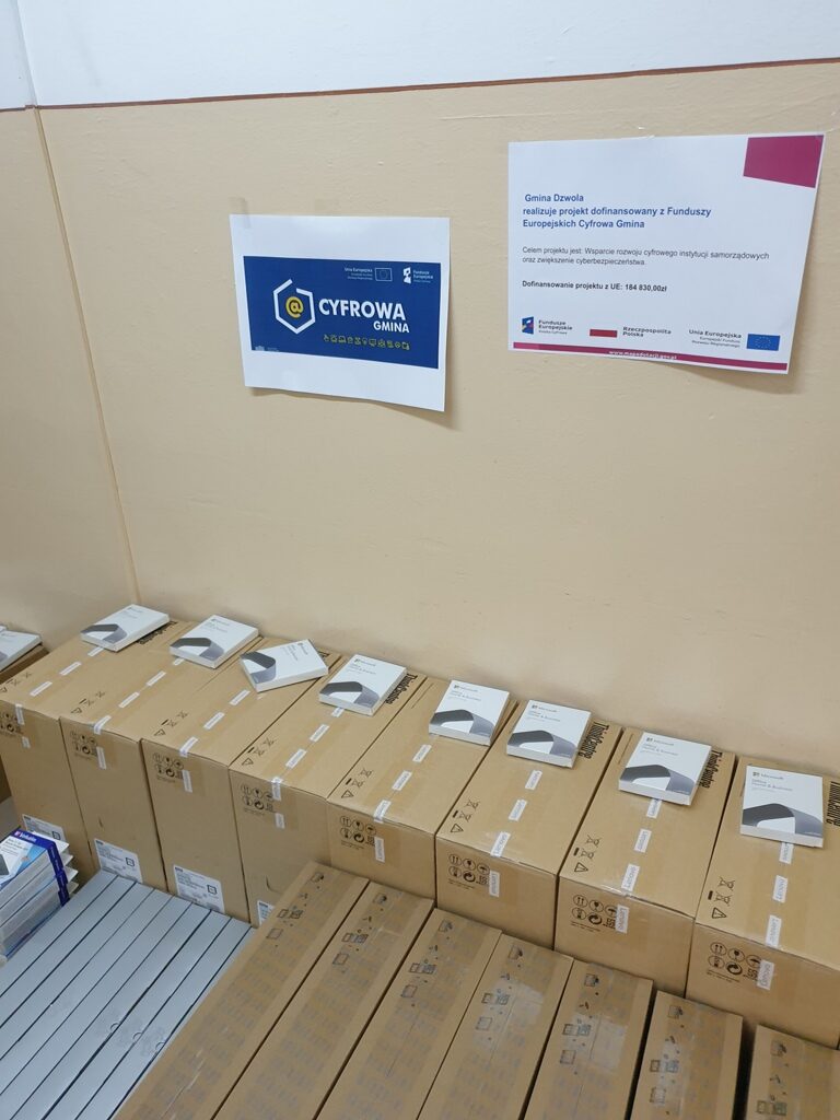 Pudełka z laptopami ustawione w rzędzie na podłodze przy beżowej ścianie z dwoma plakatami, jeden z napisem "Cyfrowa Gmina" na niebieskim tle.