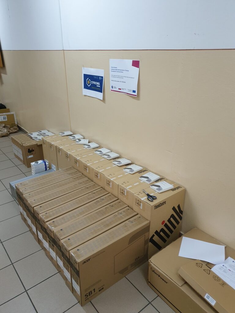 Stosy kartonowych pudełek z elektroniką ułożone przy beżowej ścianie na jasnoszarej podłodze, na pudełkach rozłożone białe instrukcje i nożyczki.