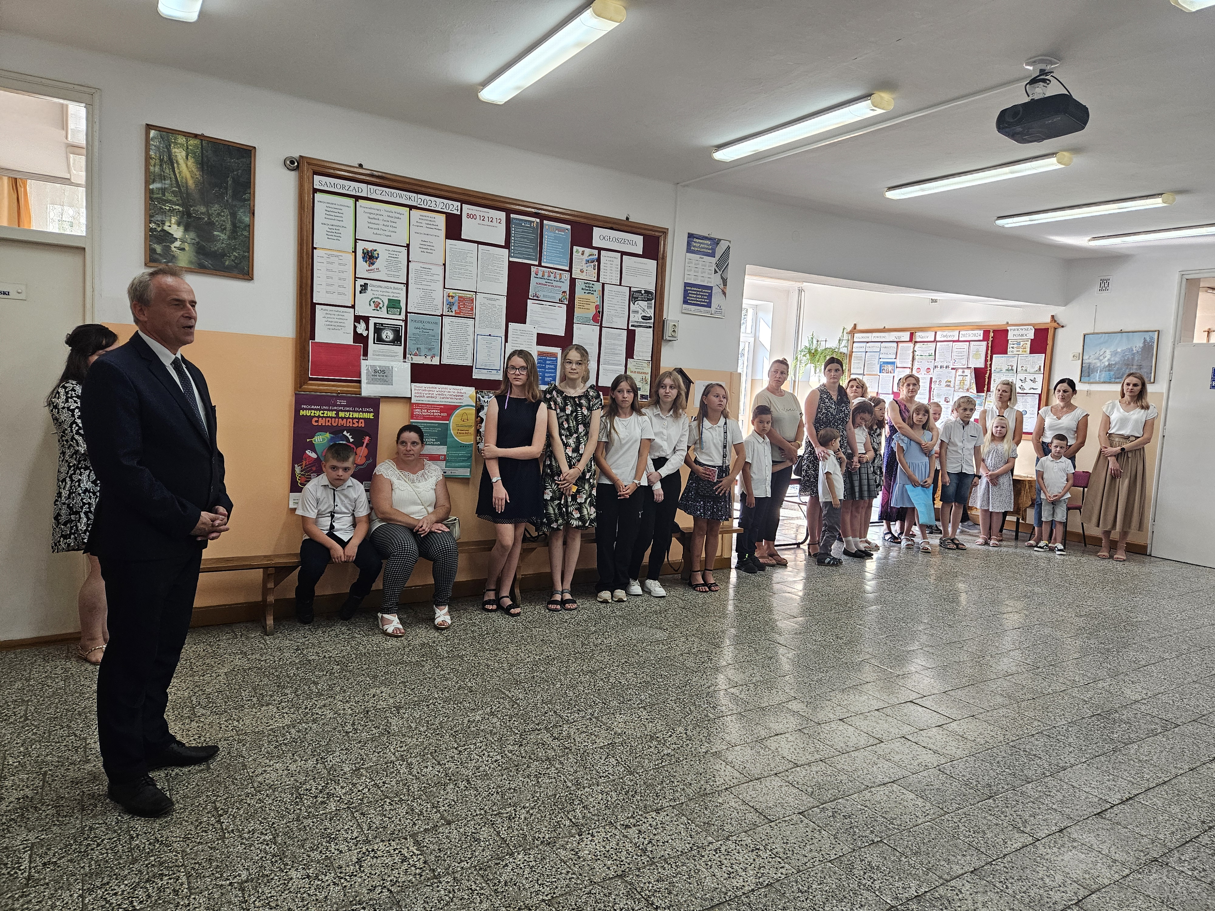 Grupa dzieci i dorosłych w jasnym korytarzu, na ścianach tablice informacyjne z licznymi ogłoszeniami, pod sufitem lampy fluorescencyjne i projektor.