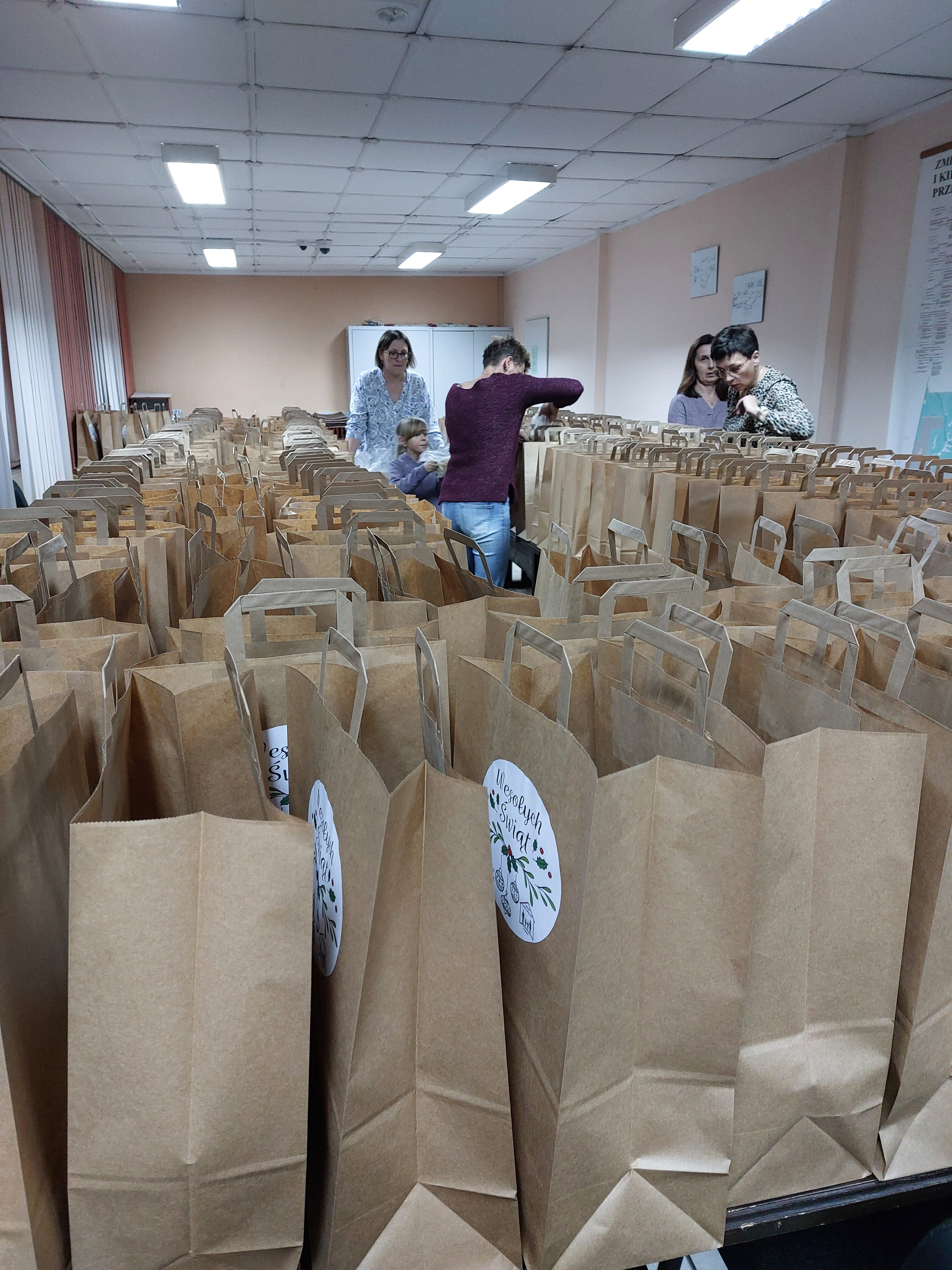 Wnętrze pomieszczenia z wieloma brązowymi papierowymi torbami z uchwytami i okrągłymi naklejkami, w tle cztery osoby przy stołach z torbami.