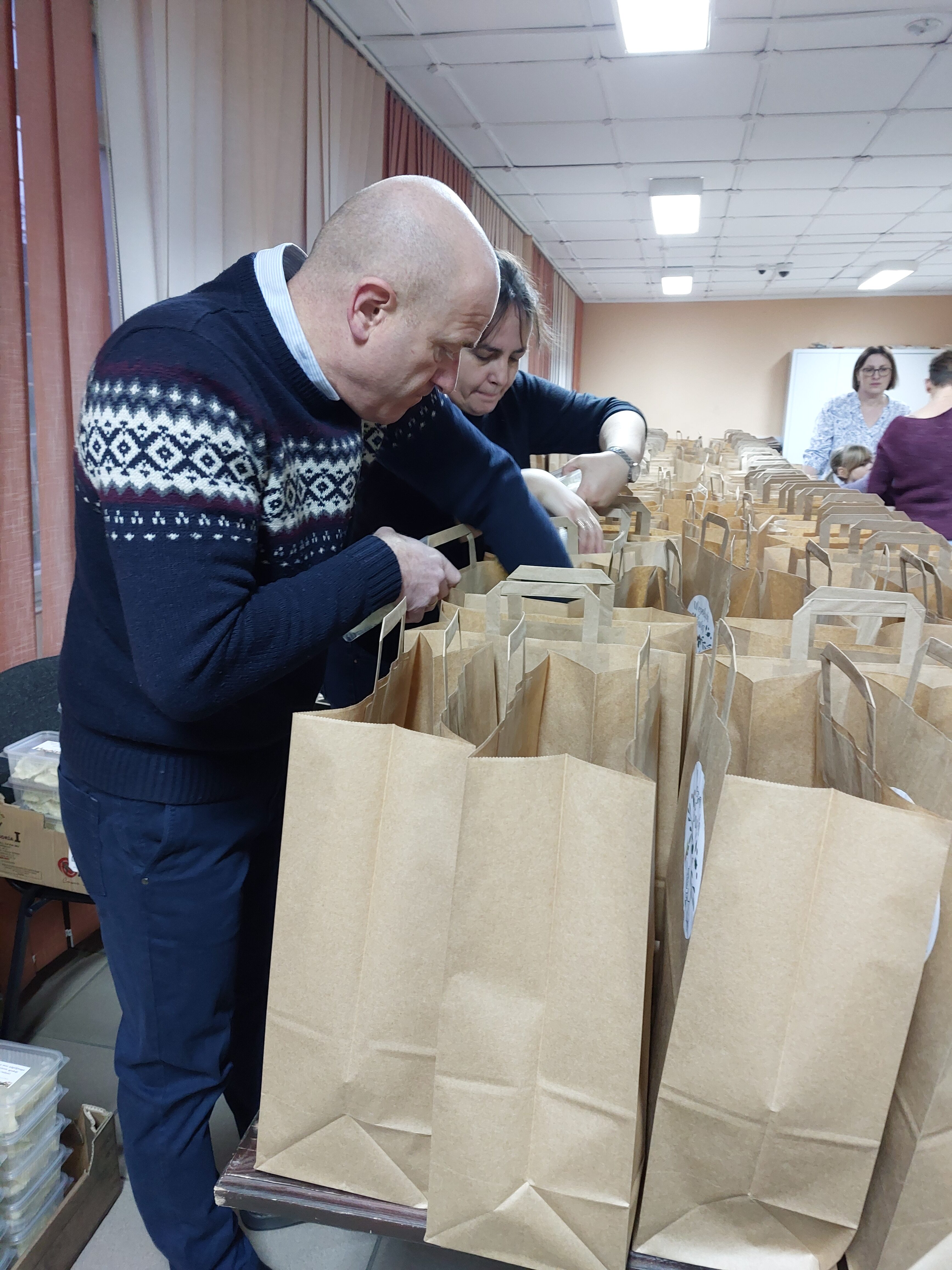Mężczyzna i kobieta pakują przedmioty do brązowych papierowych toreb na długim stole w jasnym pomieszczeniu z zasłonami i lampami sufitowymi.