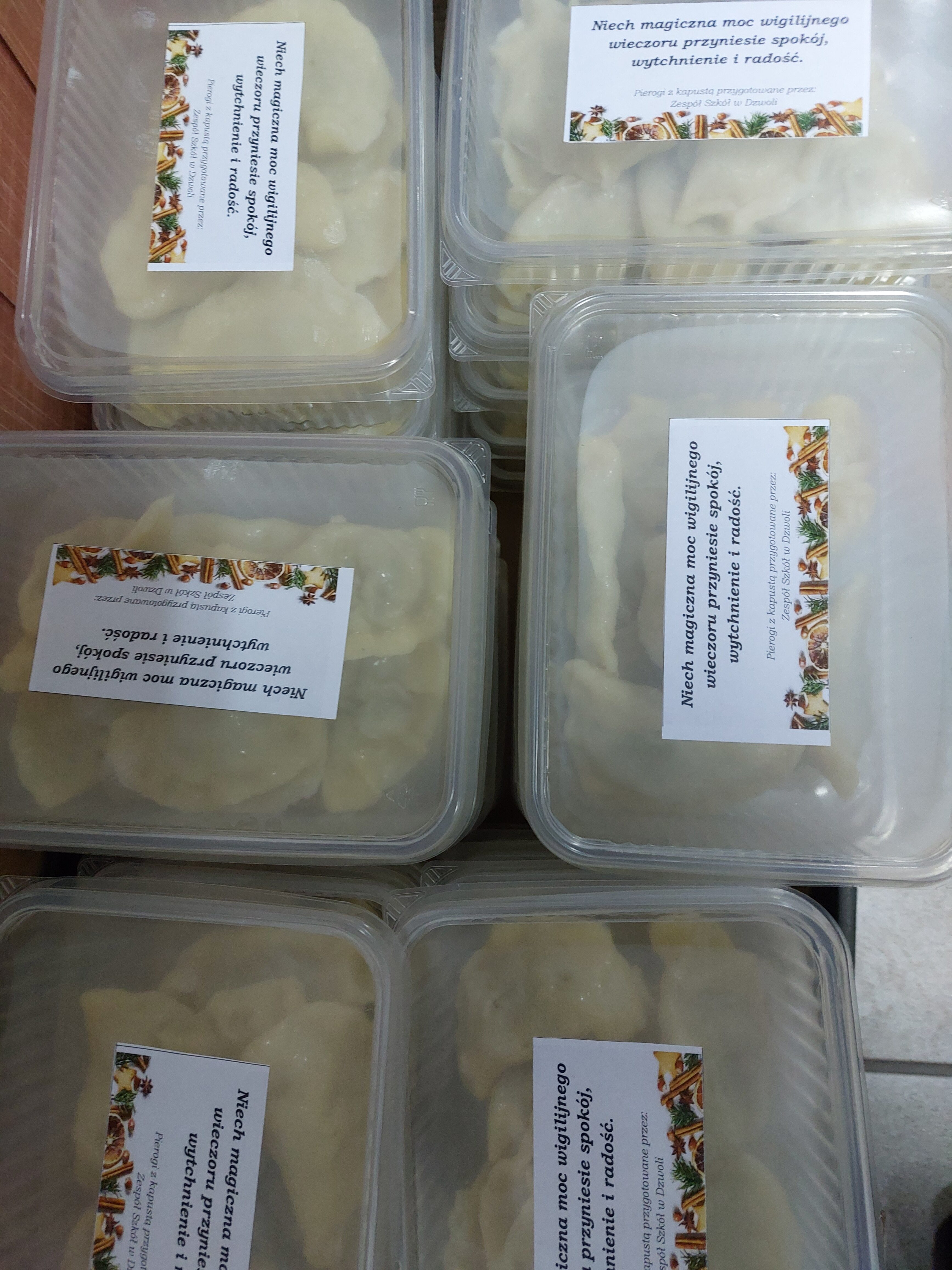 Przezroczyste plastikowe pojemniki z białymi pierogami, na każdym etykieta z napisem i ozdobnym wzorem świątecznym.