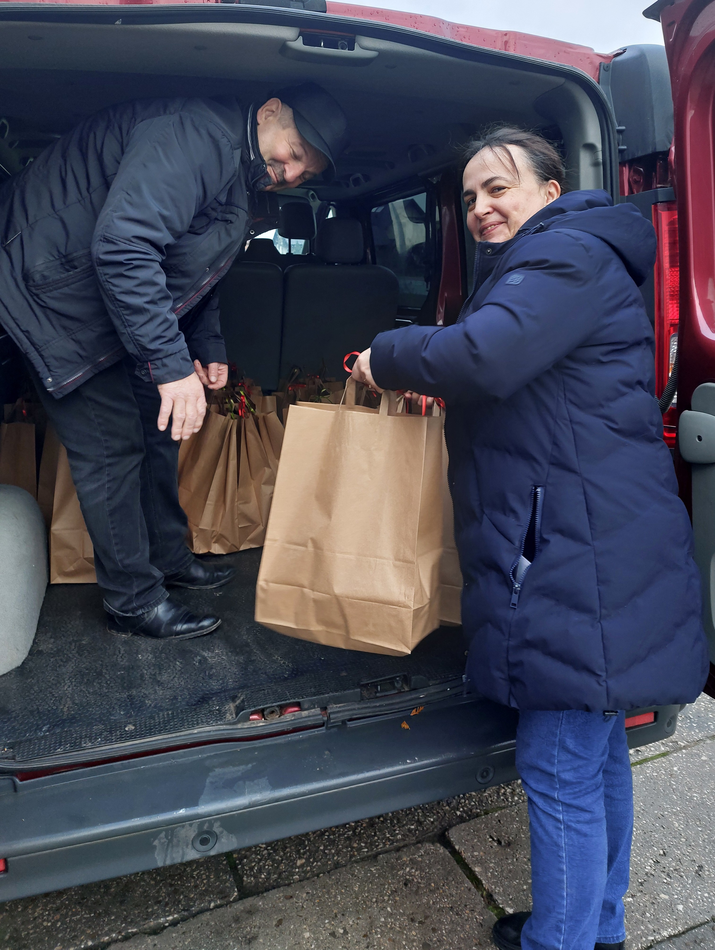 Kobieta w granatowej kurtce i mężczyzna w czarnej kurtce i czapce wyjmują papierowe torby z czerwonego pojazdu dostawczego.