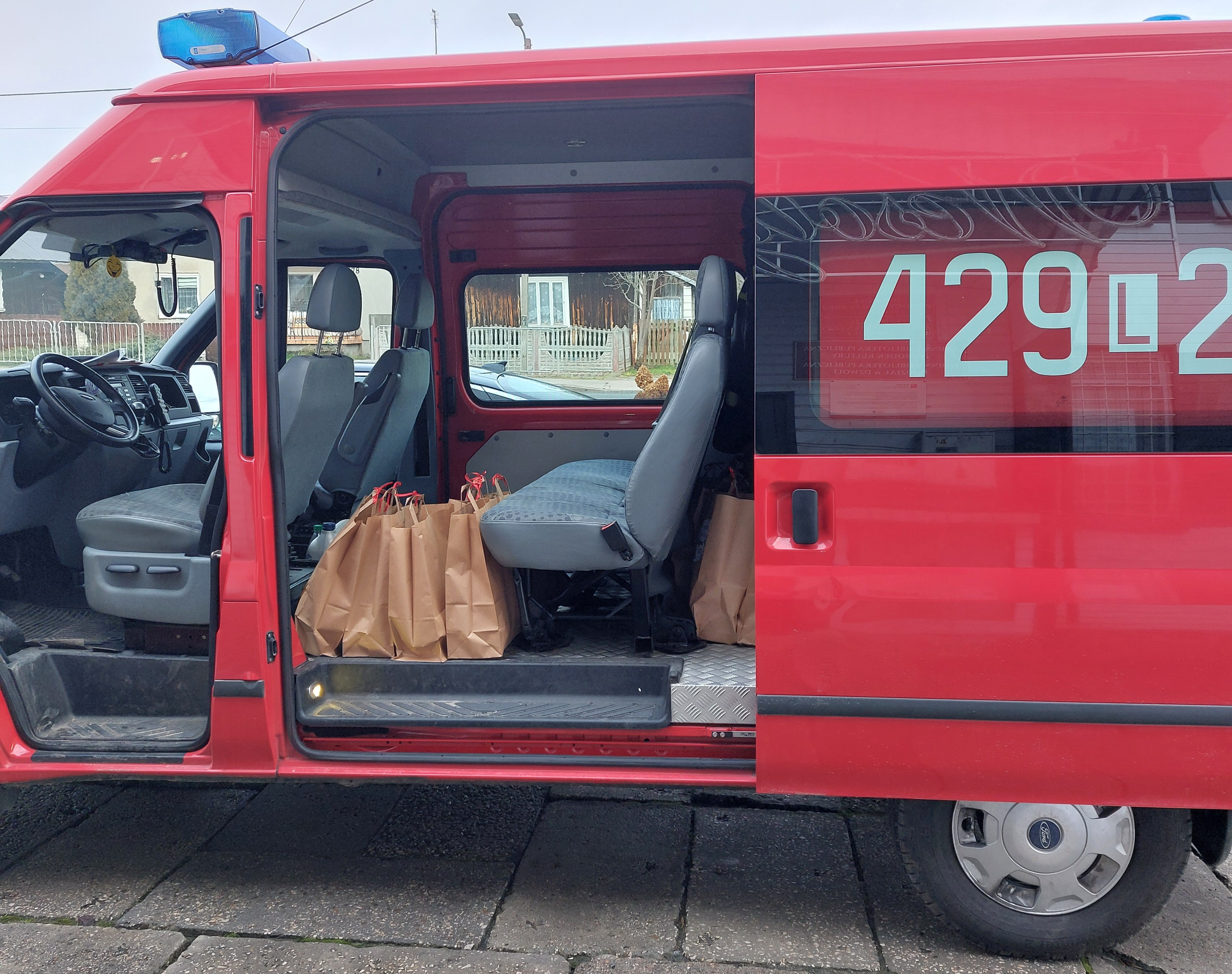 Czerwony pojazd z otwartymi drzwiami bocznymi, widoczne szare siedzenia i brązowe torby papierowe ustawione na podłodze wewnątrz. Na bocznej szybie białe cyfry.