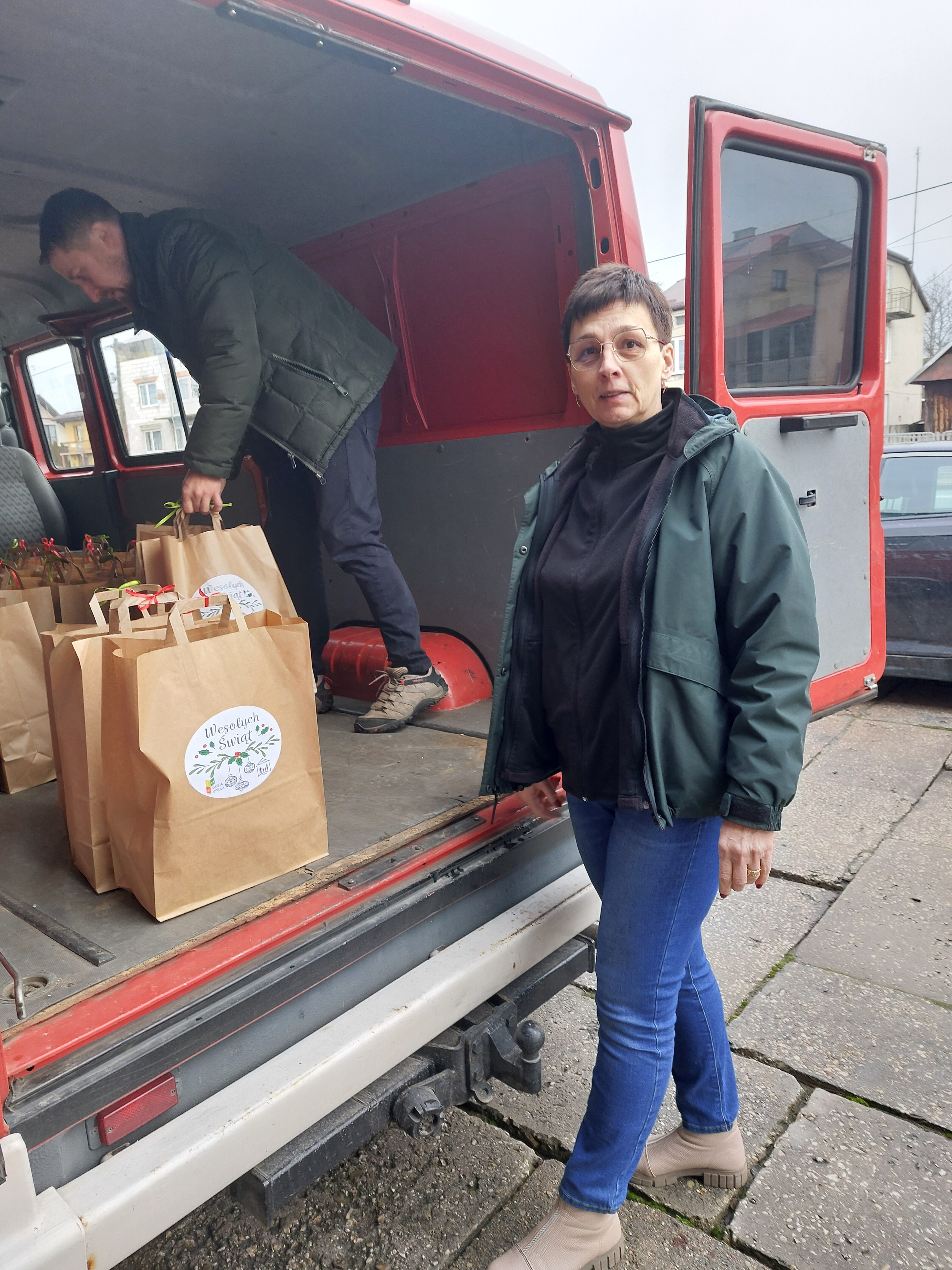 Kobieta w zielonej kurtce i niebieskich spodniach stoi obok otwartych drzwi czerwonego samochodu dostawczego, w środku mężczyzna układa papierowe torby z naklejkami "Wesołych Świąt".