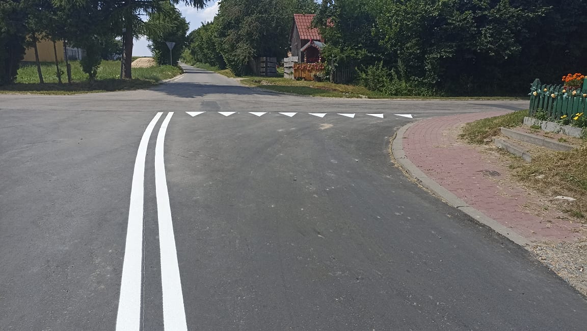 Skrzyżowanie z nową, podwójną linią ciągłą i linią zatrzymania z białych trójkątów, asfaltowa droga, z prawej strony chodnik z czerwonych kostek, zielone ogrodzenie z kwiatami.