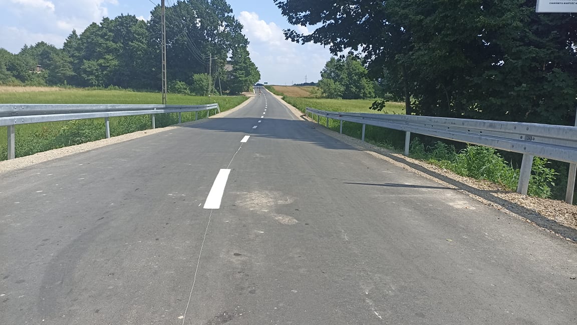 Asfaltowa droga z białym oznaczeniem na środku, osłonięta metalowymi barierkami po obu stronach, otoczona zielonymi polami i drzewami pod niebieskim niebem z chmurami.