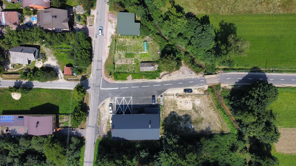 Widok z lotu ptaka na skrzyżowanie dróg, domy z dachami w kolorach szarym i brązowym, zielone pola i drzewa wokół.