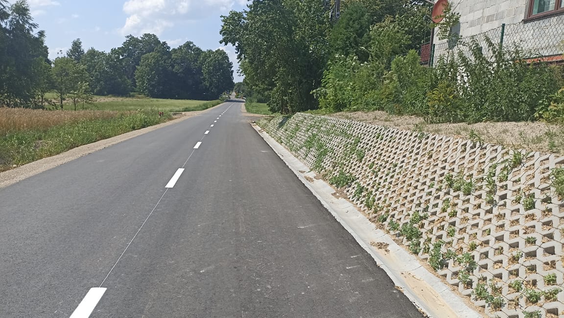 Asfaltowa droga z białymi liniami, po prawej schodkowa betonowa skarpa z roślinami, z tyłu zarośla i fragment budynku z szarą elewacją i brązowym oknem.