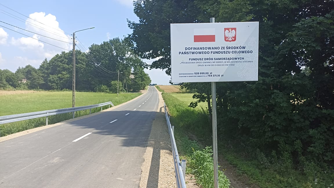 Tablica z napisem o dofinansowaniu drogi z funduszu państwowego, na tle asfaltowej drogi, zielonych pól i drzew, błękitne niebo z chmurami.