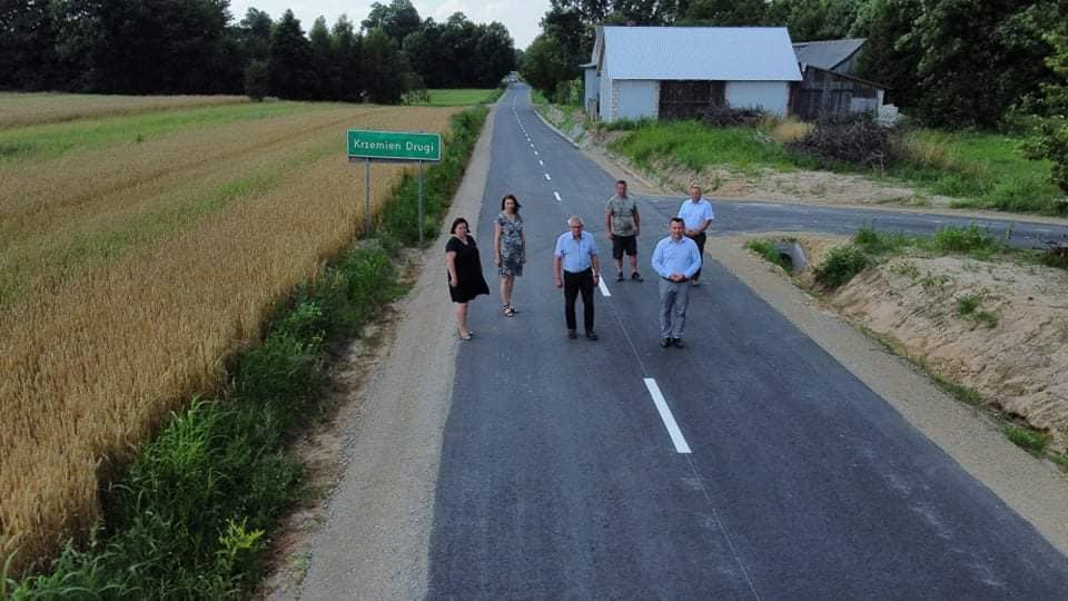 Sześć osób stoi na asfaltowej drodze z białymi liniami pośrodku, z lewej strony pole zboża, z prawej budynek z jasnym dachem i zielony znak z napisem "Kremien Drugi".