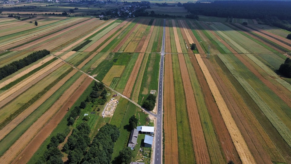Pola uprawne w różnych odcieniach zieleni, brązu i żółci, droga przecinająca pola, zalesione fragmenty oraz kilka budynków.