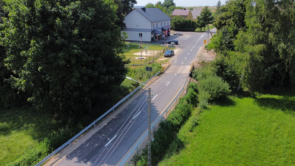 Most i droga otoczone zieloną roślinnością, po lewej duże drzewo, po prawej łąka i krzewy, w tle budynki mieszkalne i zaparkowane samochody.