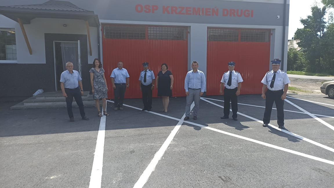 Siedem osób stoi na asfaltowym placu przed budynkiem z szarymi ścianami i czerwonymi drzwiami garażowymi, nad drzwiami widoczny napis OSP KRZEMIEŃ DRUGI.