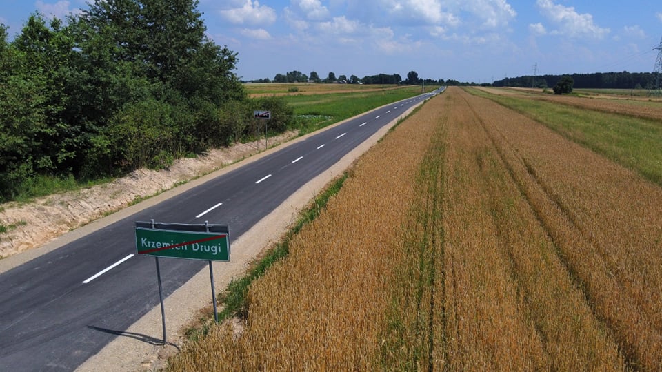 Asfaltowa droga z zielonym znakiem z przekreśloną nazwą miejscowości, po prawej stronie złote pole, po lewej zielone krzaki i drzewa pod niebieskim niebem z chmurami.
