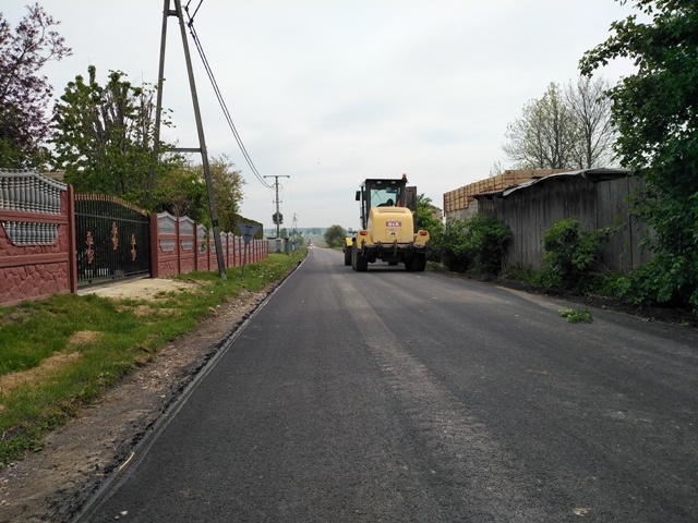 Wąska asfaltowa droga z maszyną budowlaną, po lewej stronie czerwony płot i zielona trawa, po prawej drzewa i drewniana wiata.